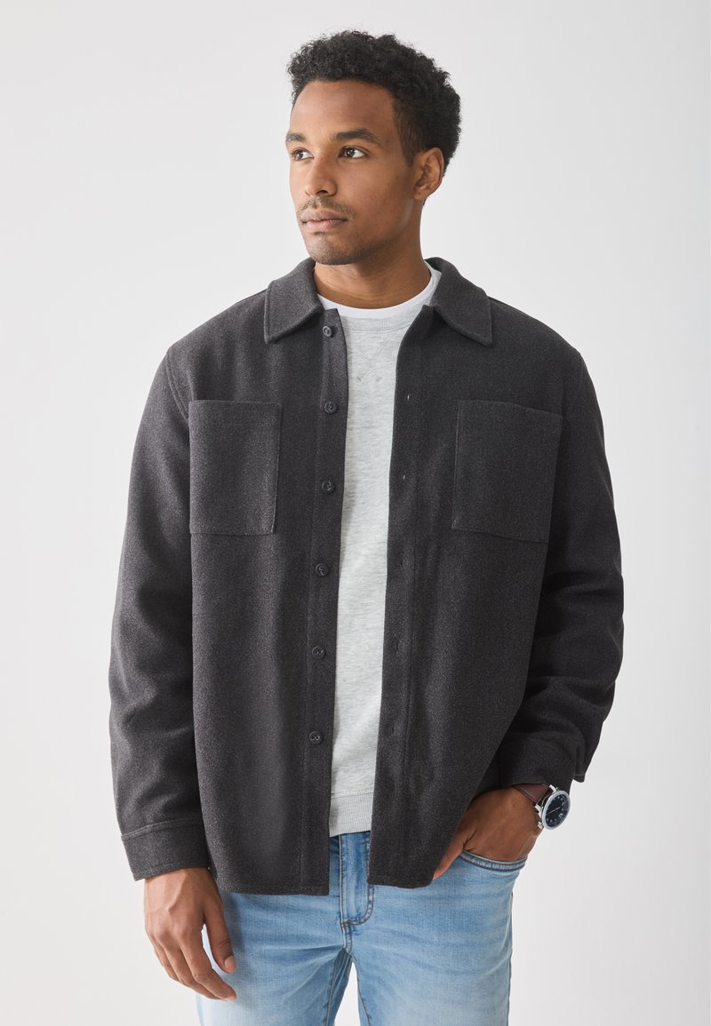 Chaqueta negra de botones con dos bolsillos frontales, hecha de tela texturizada. Combinada con una camisa gris claro y jeans azules.