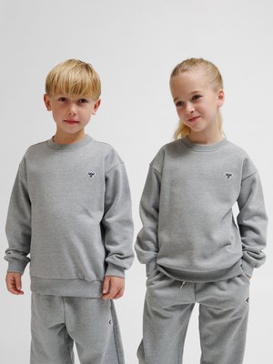 To børn iført matchende grå sweatshirts og sweatpants står side om side mod en ensfarvet baggrund.