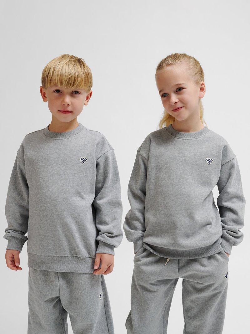 To børn iført matchende grå sweatshirts og sweatpants står side om side mod en ensfarvet baggrund.