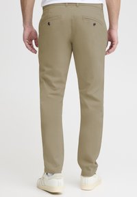 Beige trousers with a slim fit, featuring two back pockets with button closures, paired with white sneakers. Smooth texture and tailored design.