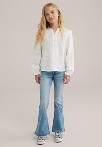 Biela košeľa na gombíky s textúrovaným, mierne zvrásneným povrchom; dlhé, vycpané rukávy; skombinovaná s svetlomodrými rozšírenými džínsami a vzorovanými teniskami.