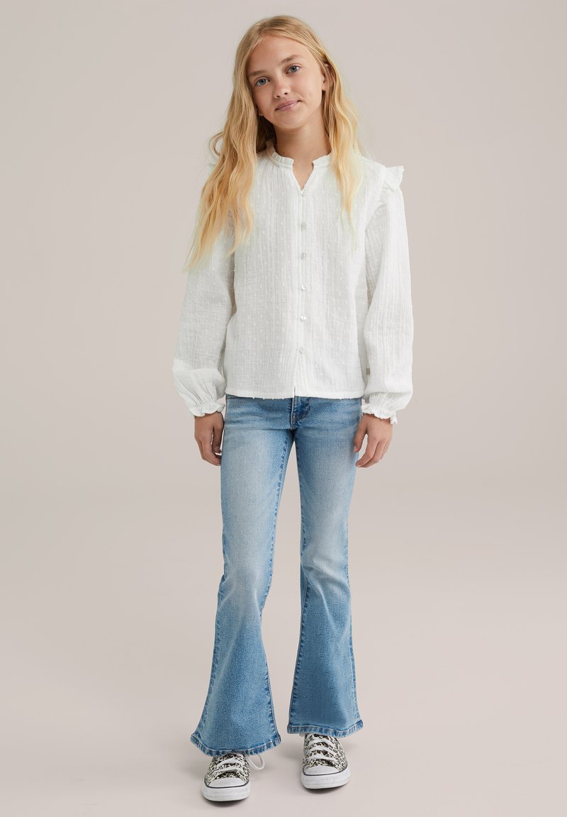 Biela košeľa na gombíky s textúrovaným, mierne zvrásneným povrchom; dlhé, vycpané rukávy; skombinovaná s svetlomodrými rozšírenými džínsami a vzorovanými teniskami.