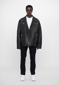 Μαύρο δερμάτινο μπουφάν μοτοσικλέτας με ασημί αξεσουάρ, που διαθέτει ασύμμετρη φερμουάρ και δύο τσέπες με φερμουάρ, φορεμένο πάνω από λευκό πουκάμισο και σκούρα τζιν.