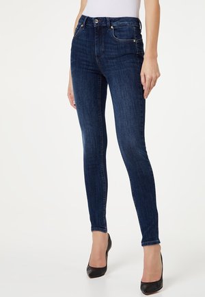 Woman wearing dark blue skinny jeans and black high heels, standing with hands relaxed by sides against a plain background.