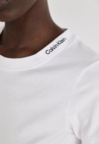 T-shirt in cotone bianco con scollo tondo, decorato con un logo "Calvin Klein" ricamato sul colletto. Texture liscia, design aderente.