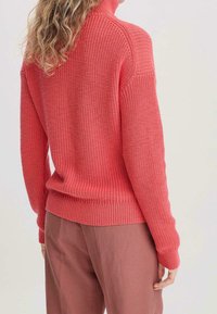 Neulottu korallinpunainen poolopaita, jossa on ribbineulos ja pitkät hihat, yhdistetty vaaleanpunaisiin housuihin, nähtynä takaa.