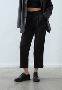 Personne portant un pantalon court noir, un crop top noir, un blazer en cuir noir et des chaussures noires à lacets, debout contre un mur clair.