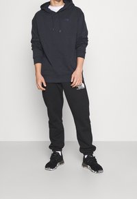 Černá mikina s kapucí a přední kapsou, s šňůrkou na stažení kapuce, kombinovaná s černými tepláky s bílým potiskem loga. Nošeno s černými teniskami.