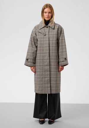 Femme debout portant un long manteau gris à carreaux avec de larges manches, un pantalon noir à jambes larges et des chaussures noires sur un fond uni.