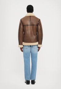 Veste en shearling marron avec un col crème, dotée d'accents en cuir et d'un design structuré. Associée à un jean bleu et des chaussures noires.