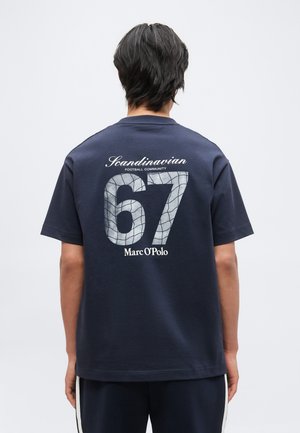 Personne vue de dos portant un T-shirt bleu marine avec "Scandinavian Football Community 67 Marc O’Polo" imprimé dans le dos.