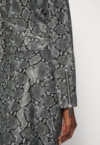 Gros plan sur le bras d'une personne portant une veste à motif peau de serpent gris et noir avec une fermeture éclair argentée sur la manche.