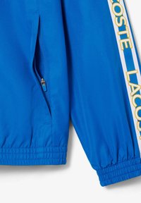 Blauwe sportjack met ritszak, elastische manchetten en een contrasterende witte streep met "LACOSTE" in gele letters. Geweven stof.