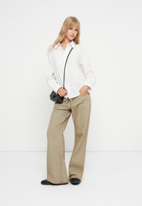 Λευκό πουκάμισο με κουμπιά, χαλαρές μπεζ παντελόνες με φράντζα στο τελείωμα και μαύρη τσάντα χιαστί. Το μοντέλο ποζάρει σε μια απλή φόντο.