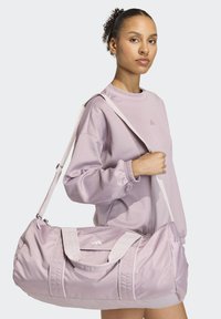 Μωβ τσάντα duffel από γυαλιστερό ύφασμα με διπλές λαβές και αποσπώμενο ιμάντα ώμου, με αντιθετικές λευκές λεπτομέρειες και φερμουάρ.