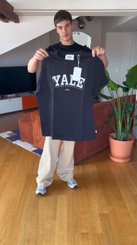 T-shirt de algodão preto com "YALE" impresso em letras maiúsculas brancas, mangas curtas, e uma etiqueta pendurada, segurado por uma pessoa contra um fundo neutro.