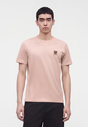Junger Mann mit kurzen Haaren, der ein schlichtes hellrosa T-Shirt mit kleinem quadratischem Logo auf der linken Brust und schwarze Hose trägt, steht vor einem schlichten Hintergrund.