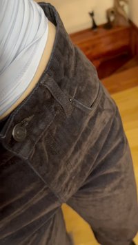 Közeli kép egy személyről, aki sötétbarna kordbársony nadrágot és fehér felsőt visel, beltéren, fa lépcső és dekoráció közelében áll.