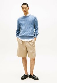 Maglione azzurro in lana a trecce, pantaloni corti beige a righe e mocassini neri; modella in piedi con le mani in tasca su uno sfondo semplice.