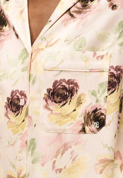 Lähedalt vaade lillemustriga särgile, millel on nööbitav tasku, suurte roosade, kollaste ja roheliste toonidega roosidega kahvatu taustal.