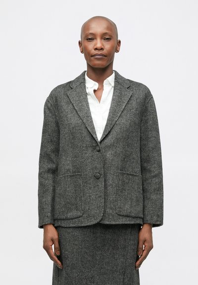 Blazer en chevrons gris avec deux poches avant et une fermeture à un bouton. Porté sur une chemise blanche à col. Fabriqué en laine texturée.