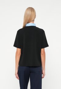 Γυναίκα με μακριά ξανθά μαλλιά που φοράει ένα μαύρο κοντομάνικο πουκάμισο πάνω από ένα ανοιχτό μπλέ πουκάμισο με γιακά και ναυτικές παντελόνες, γυρισμένη μακριά από την κάμερα.