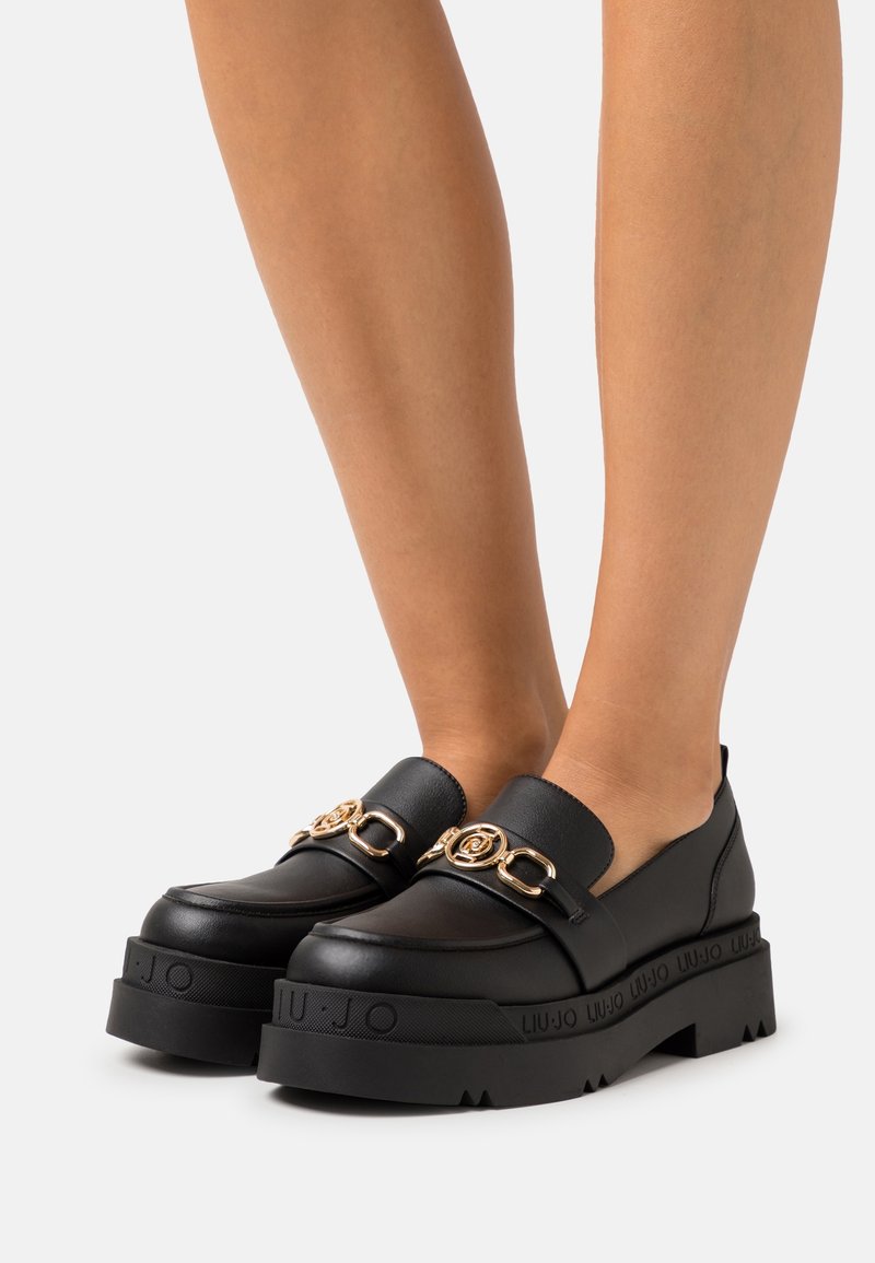 Mocassins à plateforme noire avec détail de chaîne dorée portés pieds nus sur un fond neutre.