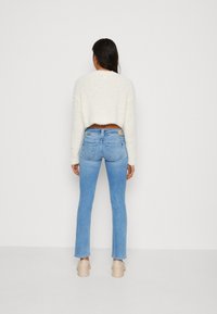 Žena stojí čelem ke zdi a má na sobě bílý zkrácený plyšový svetr, světle modré slim-fit džíny a béžové platformové boty.