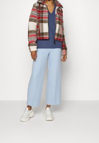 Veste en peluche courte avec un motif à carreaux rouge, gris et blanc. Pull bleu côtelé à col en V, pantalons larges bleus et baskets blanches.