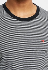 Stribet t-shirt i sort og hvid med en sort rund halsudskæring og et rødt synmaskinelogo på brystet. Fremstillet af bomuldsmateriale, afslappet pasform.