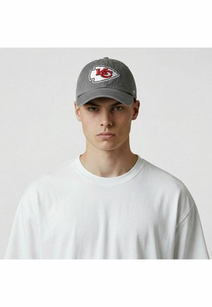 Junger Mann trägt ein schlichtes weißes T-Shirt und eine graue Baseballmütze mit dem rot-weißen Logo der Kansas City Chiefs, blickt nach vorne vor neutralem Hintergrund.