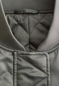 Quilted green jacket collar with ribbed knit trim and rectangular fabric tab with visible stitching on front closure.