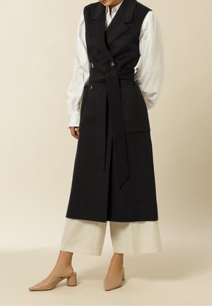 Model trägt einen langen schwarzen ärmellosen Mantel mit Gürtel und großer Tasche, eine weiße Bluse mit Puffärmeln, weite weiße Hose und beige Absatz-Mules.