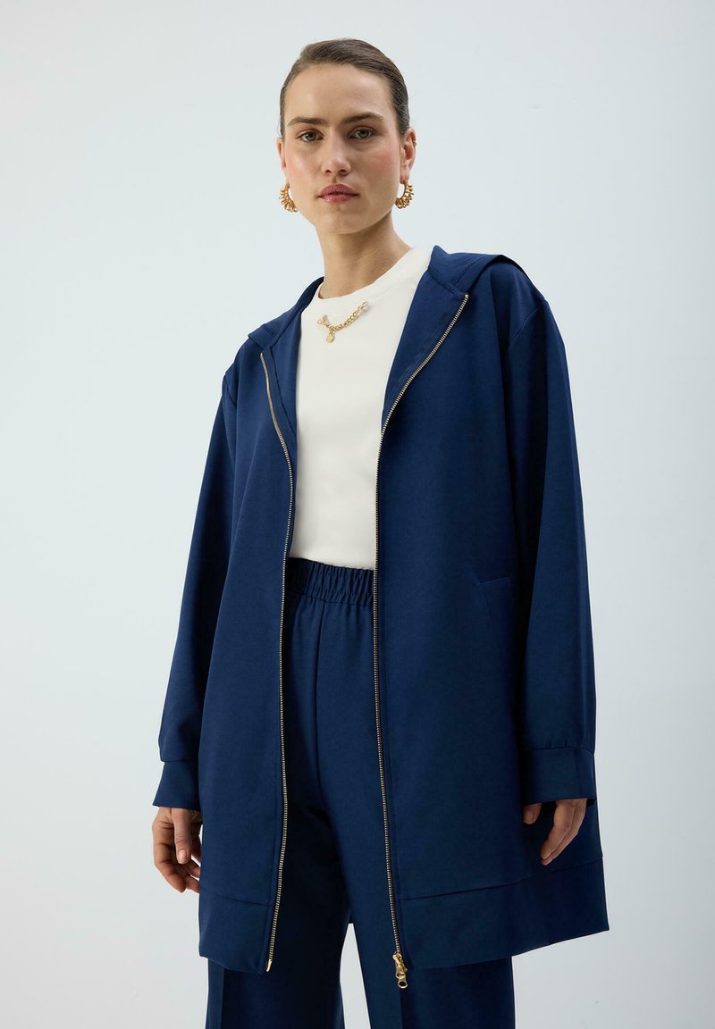 Frau trägt marineblaue, Reißverschluss-Kapuzenjacke und passende Hose mit weißem Oberteil, goldenen Creolen und goldener Halskette und steht vor neutralem Hintergrund.