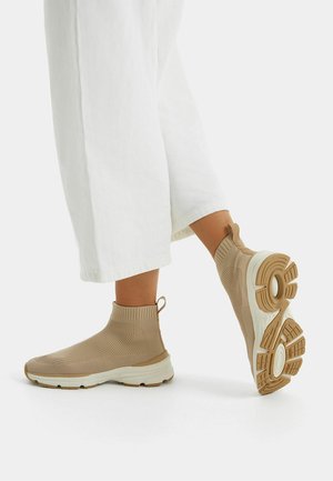 Person iført beige strikkede ankelstøvler med hvide såler og lysebrun slidbane, kombineret med hvide cropped wide-leg bukser mod en ensfarvet baggrund.