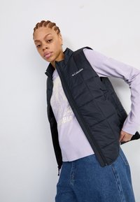 Zwarte gewatteerde vest met ritssluiting en hoge kraag, gedragen over een lichtpaarse blouse. Bevat een Columbia-logo in het wit op de borst.