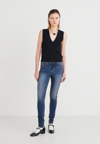 Svart ermeløs cardigan med forknapper, kombinert med blå smale jeans og svart-hvite tofargede sko. Minimalistisk design og passform.