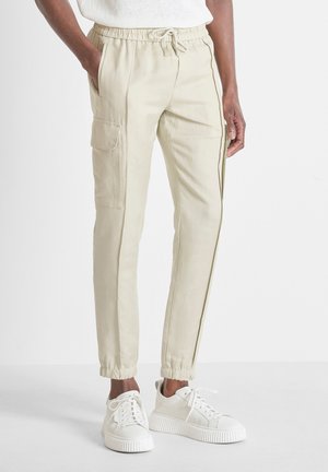 Persoon draagt beige cargobroek met elastische boorden en witte sneakers, staat met één hand in de zak tegen een effen achtergrond.