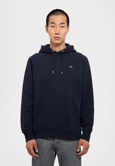 Džemperis ar kapuci tumši zilā krāsā, ar priekšējo kabatu, saites un mazu baltu logotipu. Izgatavots no mīksta auduma ar ribotām piedurknēm un apakšmalu.