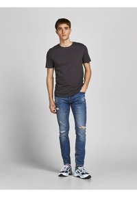 Siva bombažna majica, slim-fit modri džinsi s poškodovanimi detajli in večbarvni atletski čevlji z črnimi podplati. Preprost ozadje.