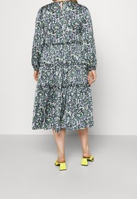 Blumenkleid mit langen Ärmeln, grüner Grundfarbe und mehrfarbigen Blumen, gestuftes Design, Rückenschlitzdetail, kombiniert mit gelben Blockabsatzsandalen.