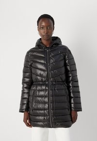 Manteau rembourré noir avec une capuche, présentant un design matelassé, une taille à cordon de serrage ajustable et des poches zippées. Fini lisse et brillant.