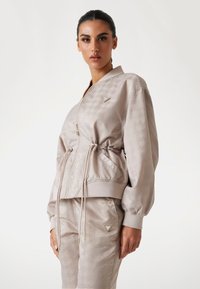 Light beige bomber jacket with a shiny finish, featuring a drawstring waist, zip closure, and embossed pattern. Gold hardware accents.