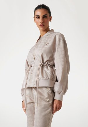 Giacca bomber color beige chiaro con finitura lucida, dotata di vita con cordoncino, chiusura con zip e motivo in rilievo. Dettagli in hardware dorato.