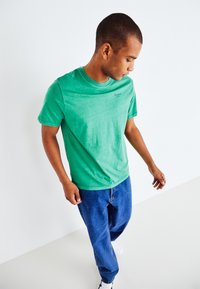 Groene katoenen t-shirt met ronde hals en korte mouwen, met een subtiele logo op de linkerborst. Draagt met blauwe denim jeans.