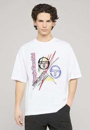Ung mand med krøllet mørkt hår iført hvid oversized T-shirt med farverigt abstrakt grafik og teksten "Sergio Tacchini", kombineret med sorte bukser.