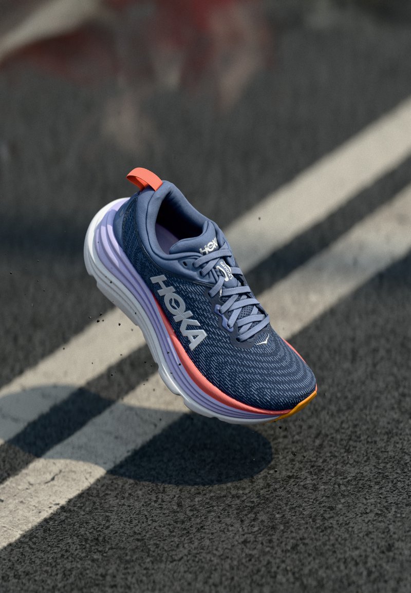 HOKA löparsko i marinblått med texturerad ovansida, vit mellansula och orange accentdetaljer. Har en lätt design och vadderad krage.