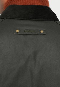 Σκούρο πράσινο μπουφάν με γιακά από κορντόρι, που διαθέτει ραμμένη ετικέτα με το όνομα της μάρκας "Barbour" και διακοσμητικό λεπτομέρεια από ορείχαλκο σε κούμπωμα.