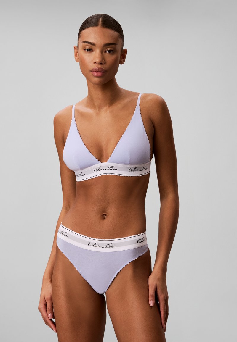 Γυναίκα που φοράει ανοιχτό μωβ bralette Calvin Klein και ταιριαστό εσώρουχο, στέκεται μπροστά σε ένα απλό γκρι φόντο.