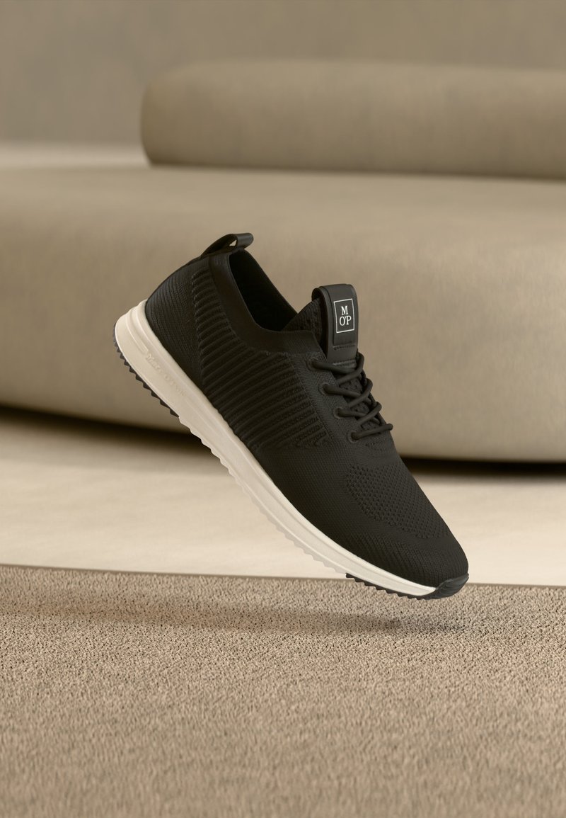 Baskets en maille noires avec semelle blanche et étiquette "MOP" sur la languette, positionnées sur un tapis beige devant un canapé beige.